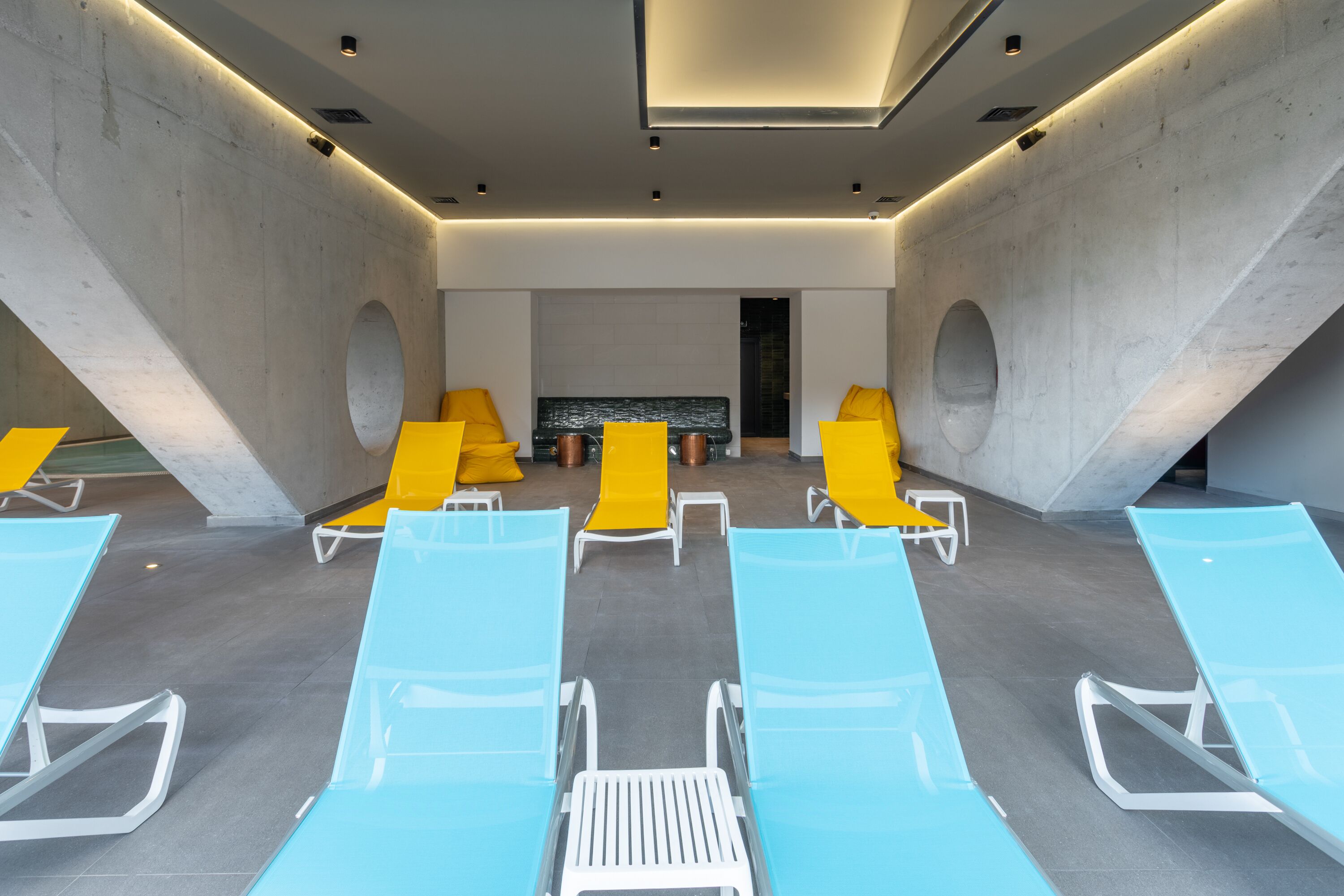 A modern indoor pool area with blue and yellow lounge chairs, a stone wall, and a central seating area with a black bench.