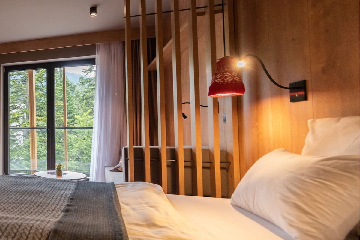 Een moderne hotelkamer met een groot bed, een nachtkastje, een lamp en een raam met uitzicht op bomen.