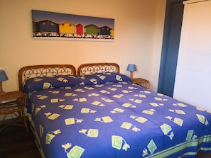 Cama doble con colcha azul con motivos geométricos, cabecera de madera, mesita de noche con lámpara y un cuadro colgado en la pared que representa cabañas de playa coloridas.