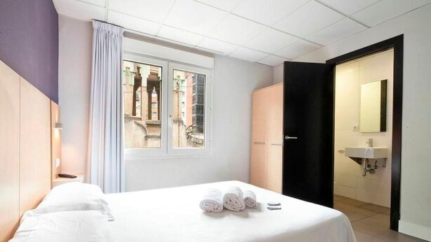 Habitación de hotel con cama, ventana con cortinas, baño con lavamanos y espejo, y vistas a edificios.