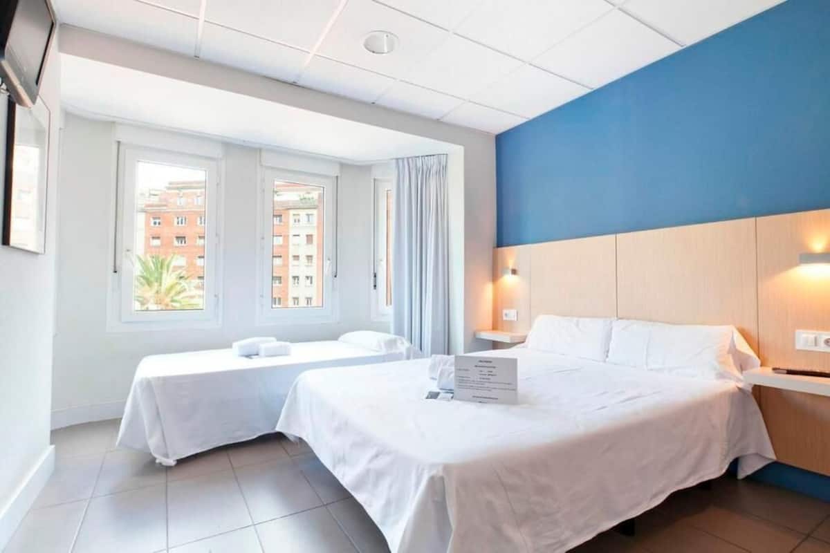 Habitación de hotel con dos camas, un ventanal con cortinas, pared de acento azul y vista a edificios.