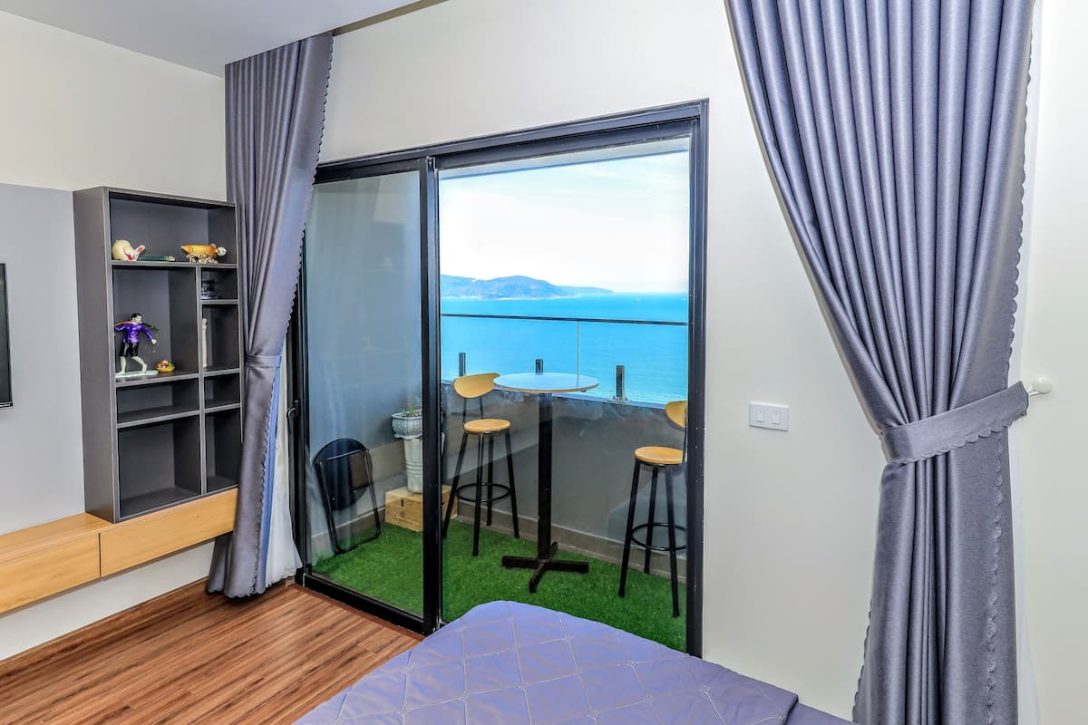 A room with a bed, a small table with chairs, a bookshelf, and a balcony with a view of the sea.