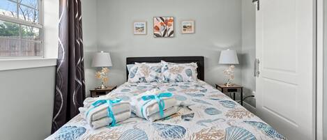 Cama bem arrumada com colcha estampada com conchas, duas luminárias de cabeceira e uma janela com cortinas.
