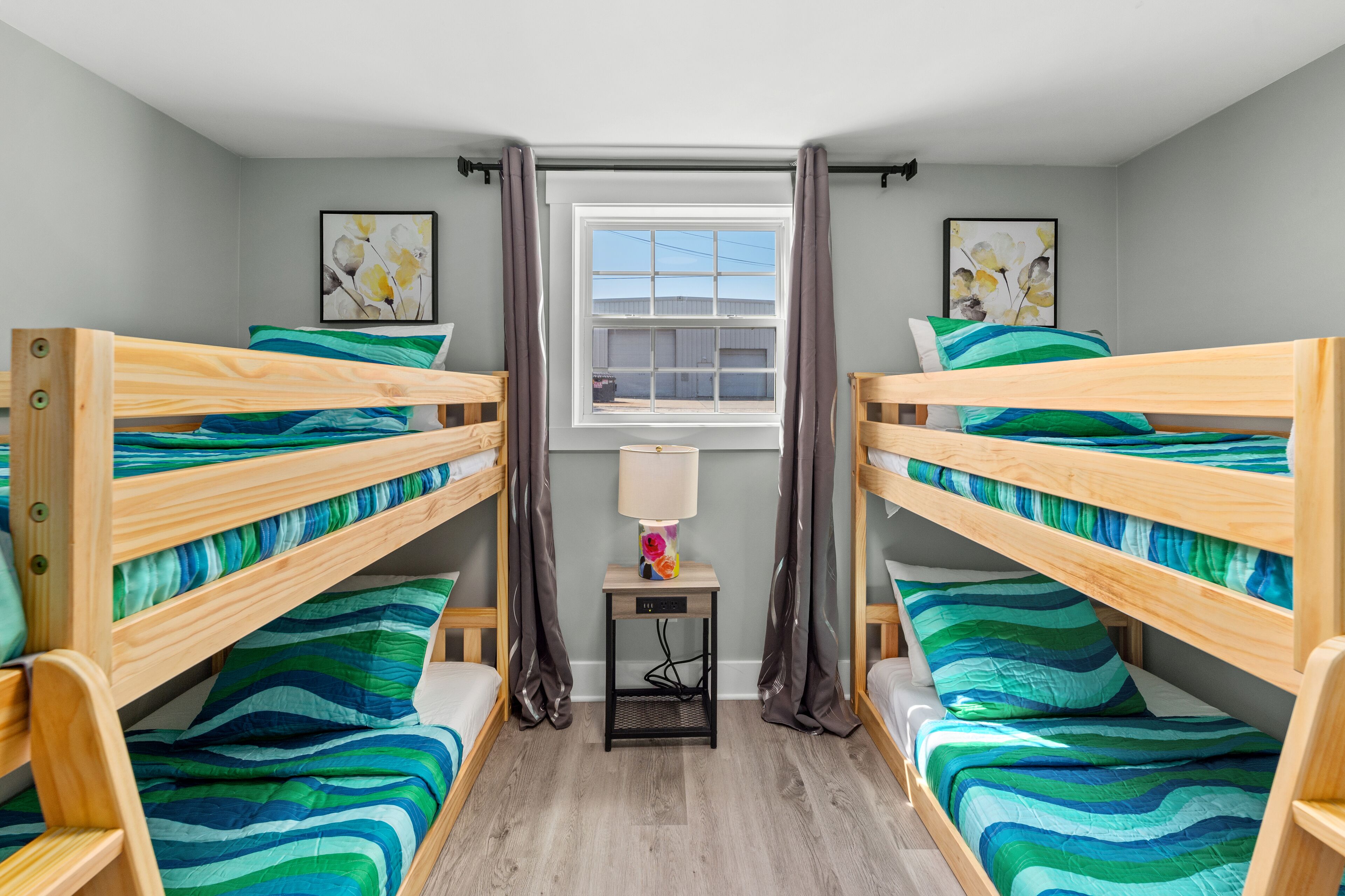 A room with three bunk beds, a small table with a lamp, and two framed pictures on the walls.