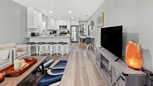 Una cocina moderna con armarios blancos, una isla central con taburetes, un comedor con mesa y sillas, un televisor de pantalla plana y una lámpara de sal.
