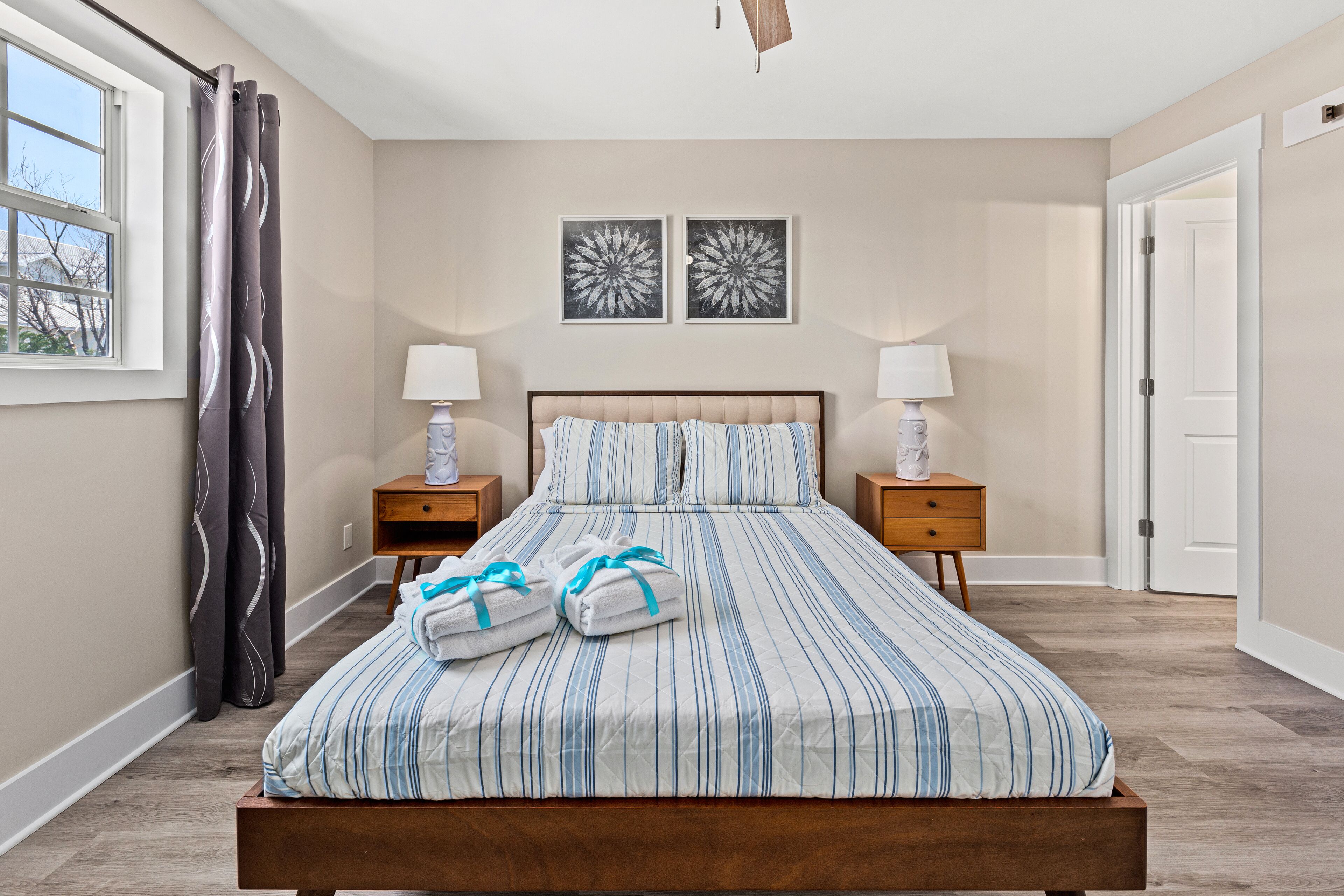 Une chambre à coucher avec un grand lit, deux tables de chevet avec des lampes, une fenêtre avec des rideaux et deux tableaux représentant des fleurs, accrochés au mur.