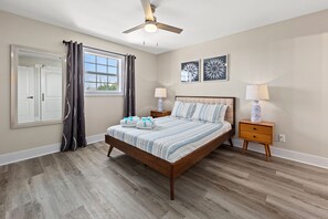 A bedroom with a wooden bed, two bedside tables, a ceiling fan, and a window with curtains. - Welcome to Dolphin Fun B, Brand NEW Duplex Walk to the Beach (Panama City Beach)