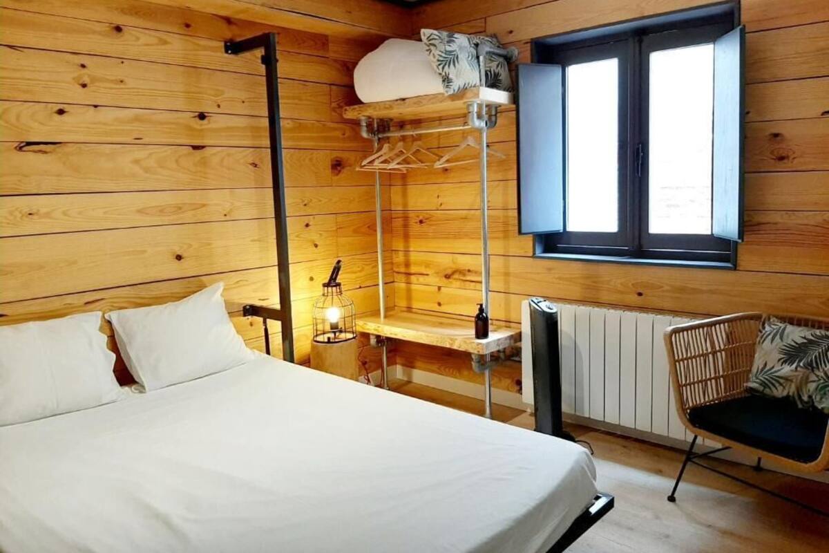 Une pièce de cabane en bois comprenant un lit, un portant à vêtements, une petite table avec une lampe et une fenêtre.