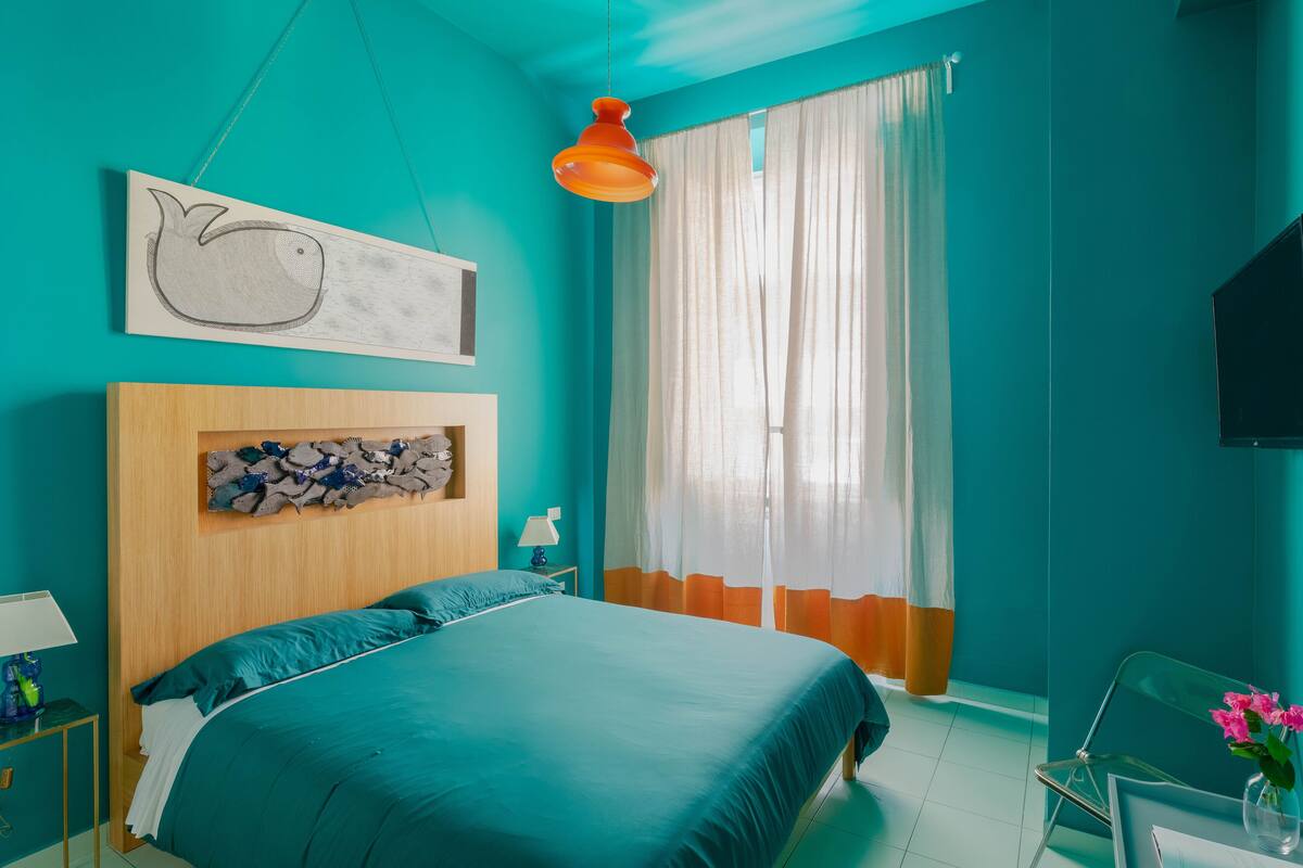 A bedroom with teal walls, a bed with teal bedding, a wooden headboard, a wall-mounted TV, a window with sheer curtains, and a hanging orange pendant light.