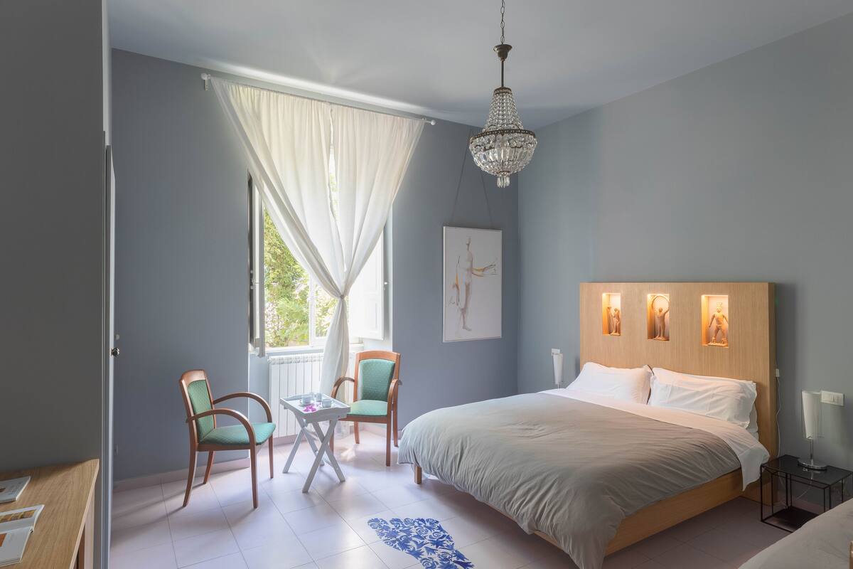 A bedroom with a large bed, a chandelier, a window with curtains, and a chair.