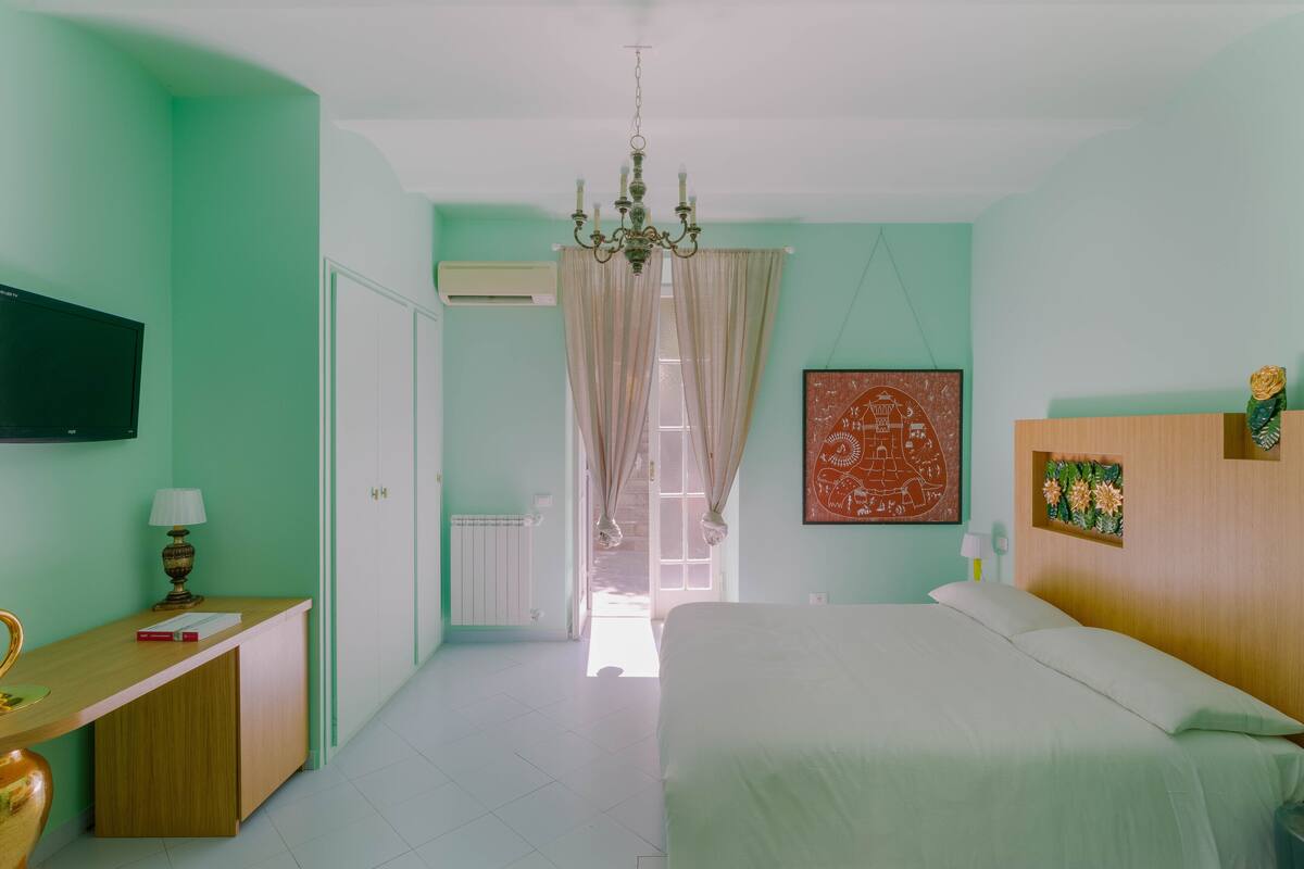 A bedroom with a bed, a desk, a chair, a television, and a chandelier.