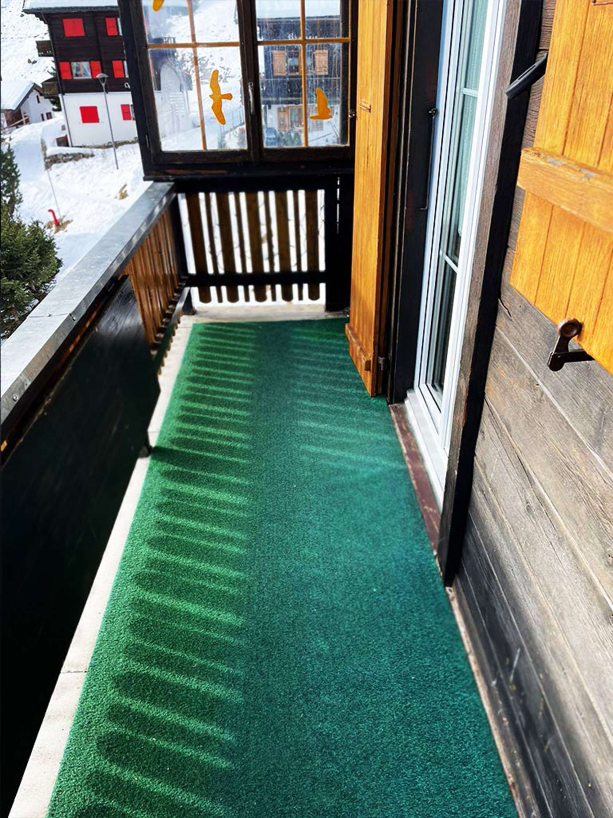 Un étroit couloir extérieur, recouvert d’un gazon synthétique vert, mène à une porte en bois.