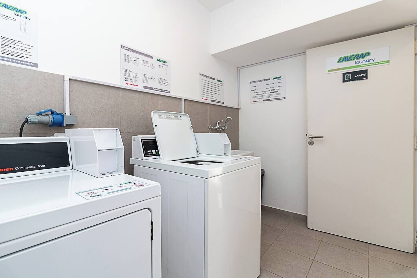 A laundry facility with commercial dryers and washing machines, a water fountain, and informational posters on the walls.