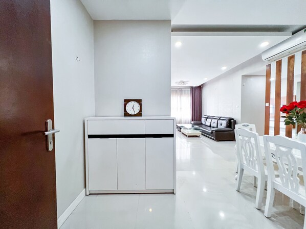 Uma sala de estar moderna com um sofá branco, uma mesa de centro e um espaço de refeições com cadeiras brancas. Um relógio está fixado na parede, e uma porta com moldura marrom é visível à esquerda.