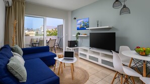 Een moderne woonkamer met een blauwe bank, een televisie, een laptop en een eethoek met een fruitschaal.
