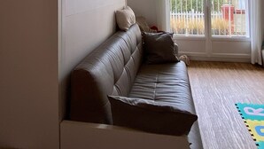 A living room with a brown leather sofa, a white wardrobe, and a large sliding glass door leading to a balcony with a view of buildings and a fence. - Calm sea view family appartment in historical town center (Villers-sur-Mer)