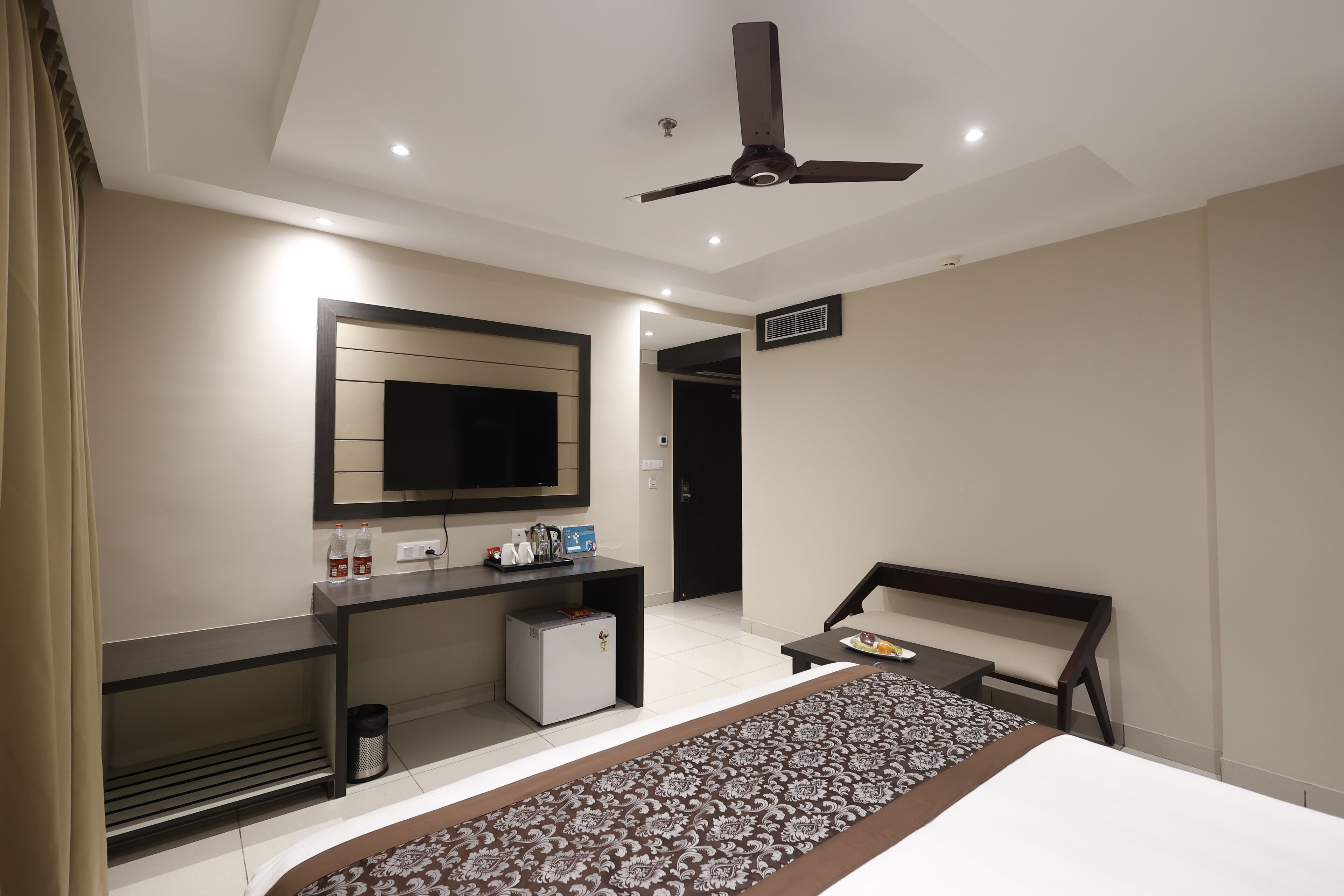 A modern hotel room with a bed, a desk with a flat-screen TV, a small refrigerator, a ceiling fan, and a bathroom door.