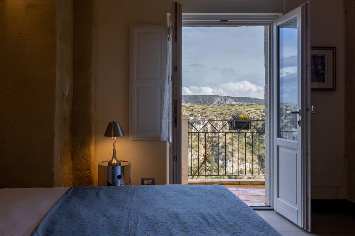 A bedroom with a bed, a nightstand, a lamp, and a balcony door leading to a scenic view.