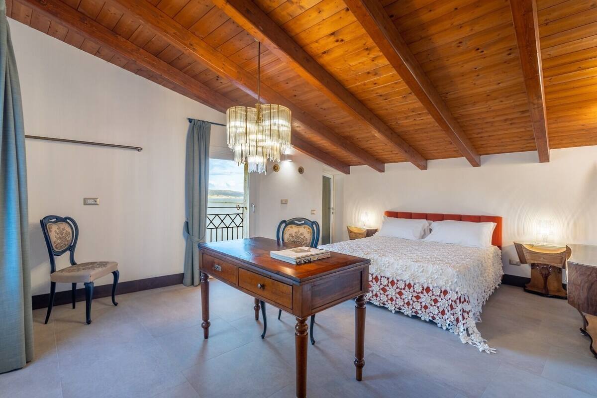 A bedroom with a wooden ceiling, a bed with white bedding, a desk with a chair, and a chandelier.