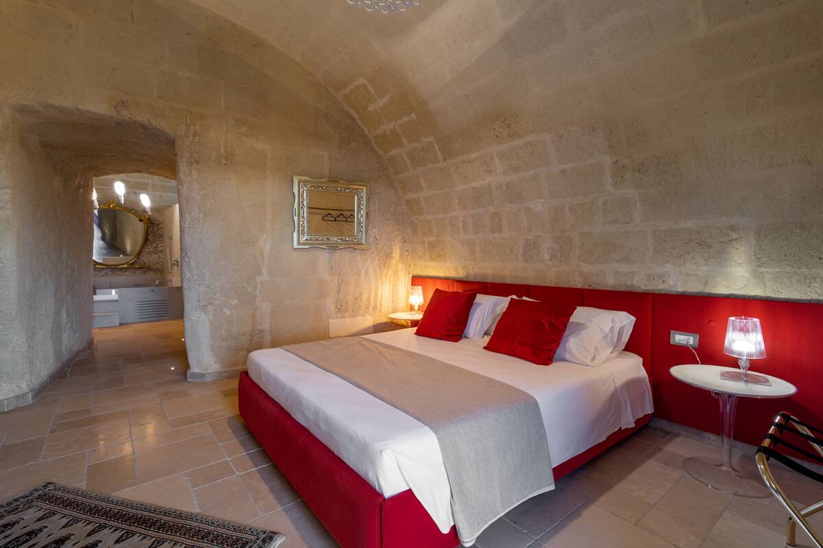 A bedroom with a red headboard, a bed with white and red bedding, a small round table with a lamp, and a mirror on the wall.