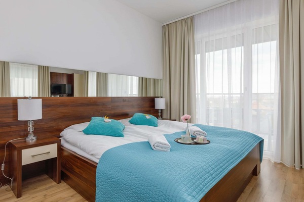 A cozy bedroom featuring a double bed with blue bedding, bedside tables, and large windows with white curtains.