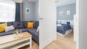 A modern room with a grey sofa, a wooden coffee table, a bed with a blue bedspread, and a white door. - Spacious Apartment | Balcony, Parking, 400 m to the Beach (Grzybowo)