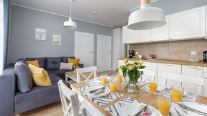 A modern living room with a grey sofa, white dining table set with plates and glasses, and a kitchen area with white cabinets and a wooden countertop. - Spacious Apartment | Balcony, Parking, 400 m to the Beach (Grzybowo)