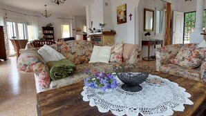 Un salón amplio con un sofá grande estampado con flores, una mesa de centro de madera con un mantel de encaje, y vistas al área de cocina y comedor a través de un arco abierto.