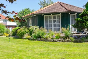 Une maison aux murs peints en vert, dotée d’un toit recouvert de tuiles et de grandes fenêtres, entourée d’un jardin bien entretenu.