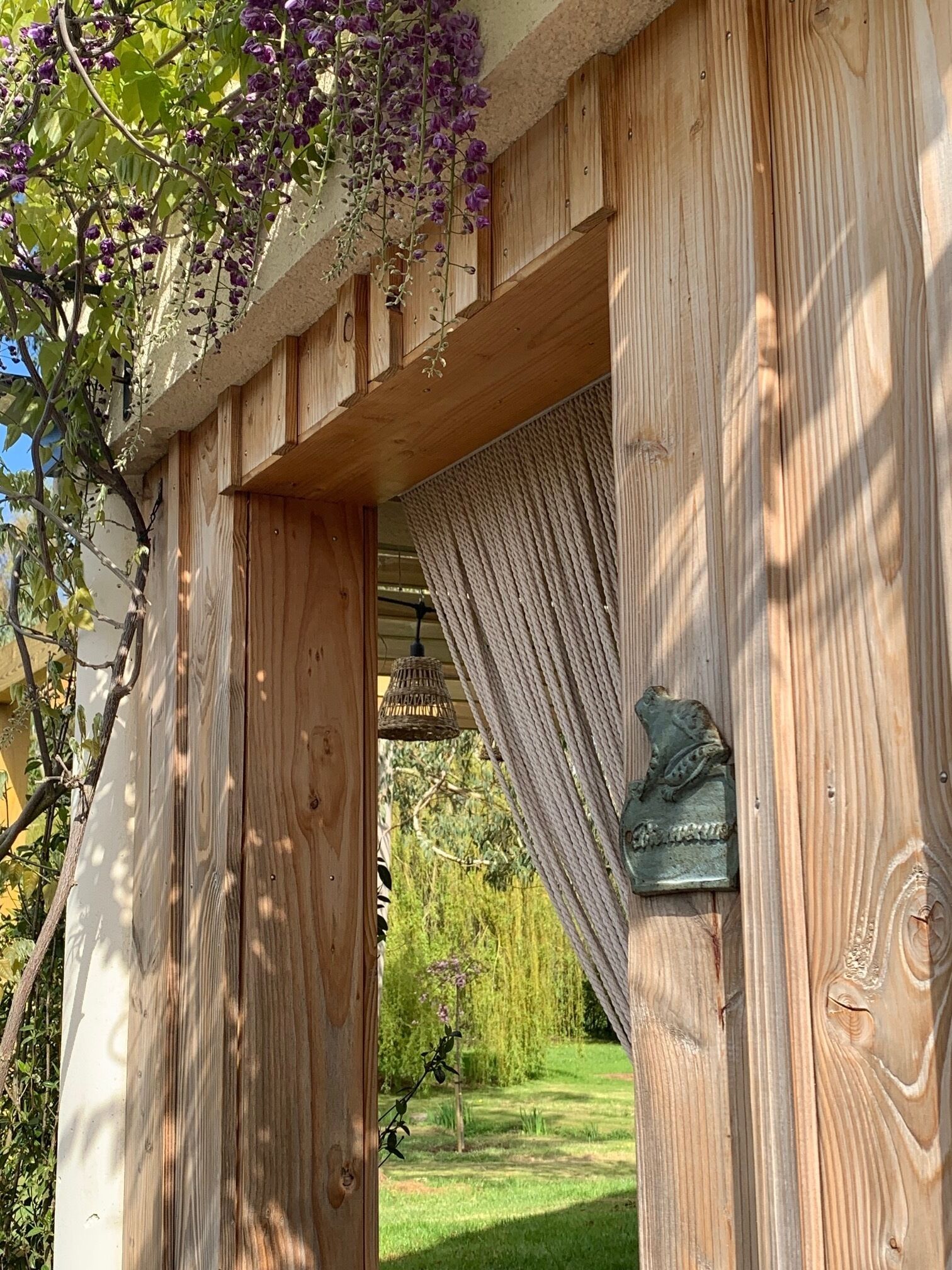 Eine Pergola aus Holz mit einem gewebten Baldachin, einer Metallschild und Blick auf einen Garten.