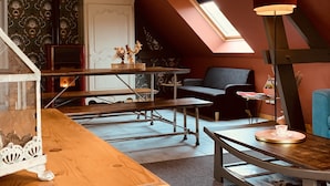 Une chambre mansardée chaleureuse, avec un plafond en pente, une lucarne, un bureau en bois, une chaise et une petite table sur laquelle se trouve une tasse.