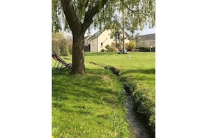Eine Wiese mit einem Baum, einem kleinen Bach und einem Gebäude im Hintergrund.