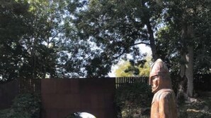 Une statue en bois représentant une figure vêtue d’une tenue traditionnelle, posée sur un socle en bois, avec un pont de pierre et de la verdure en arrière-plan.