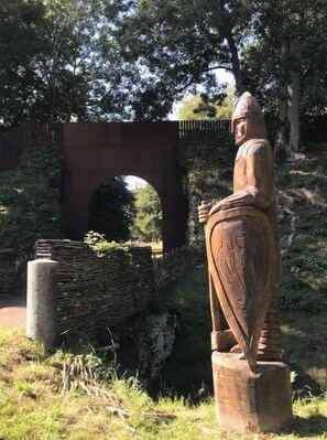 Eine hölzerne Statue einer Person in traditioneller Kleidung, stehend auf einem Holzfuß, mit einer Steinbrücke und Grünpflanzen im Hintergrund.