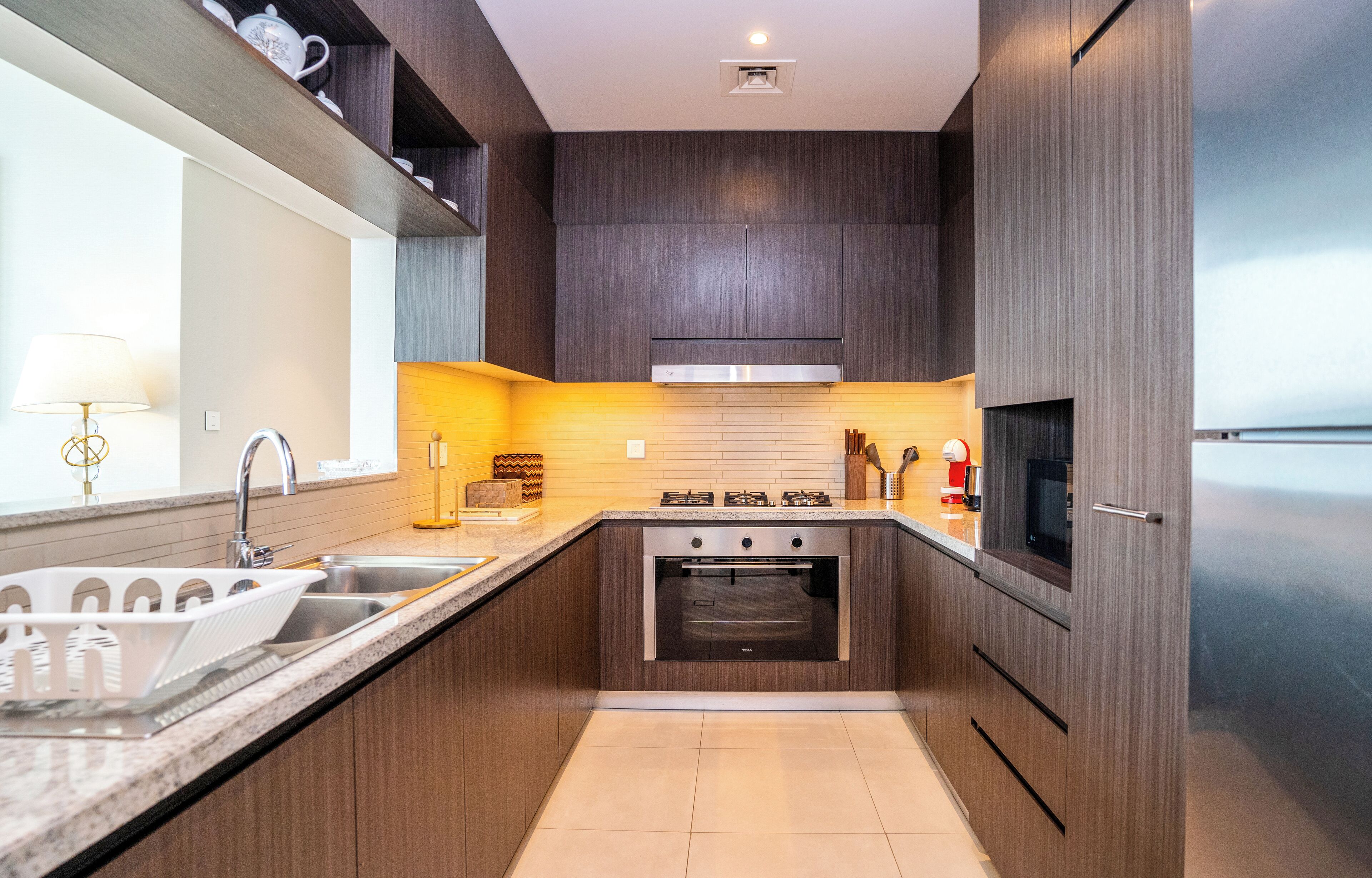 A modern kitchen with dark wood cabinets, a stainless steel range hood, a built-in oven, and a large window providing natural light.