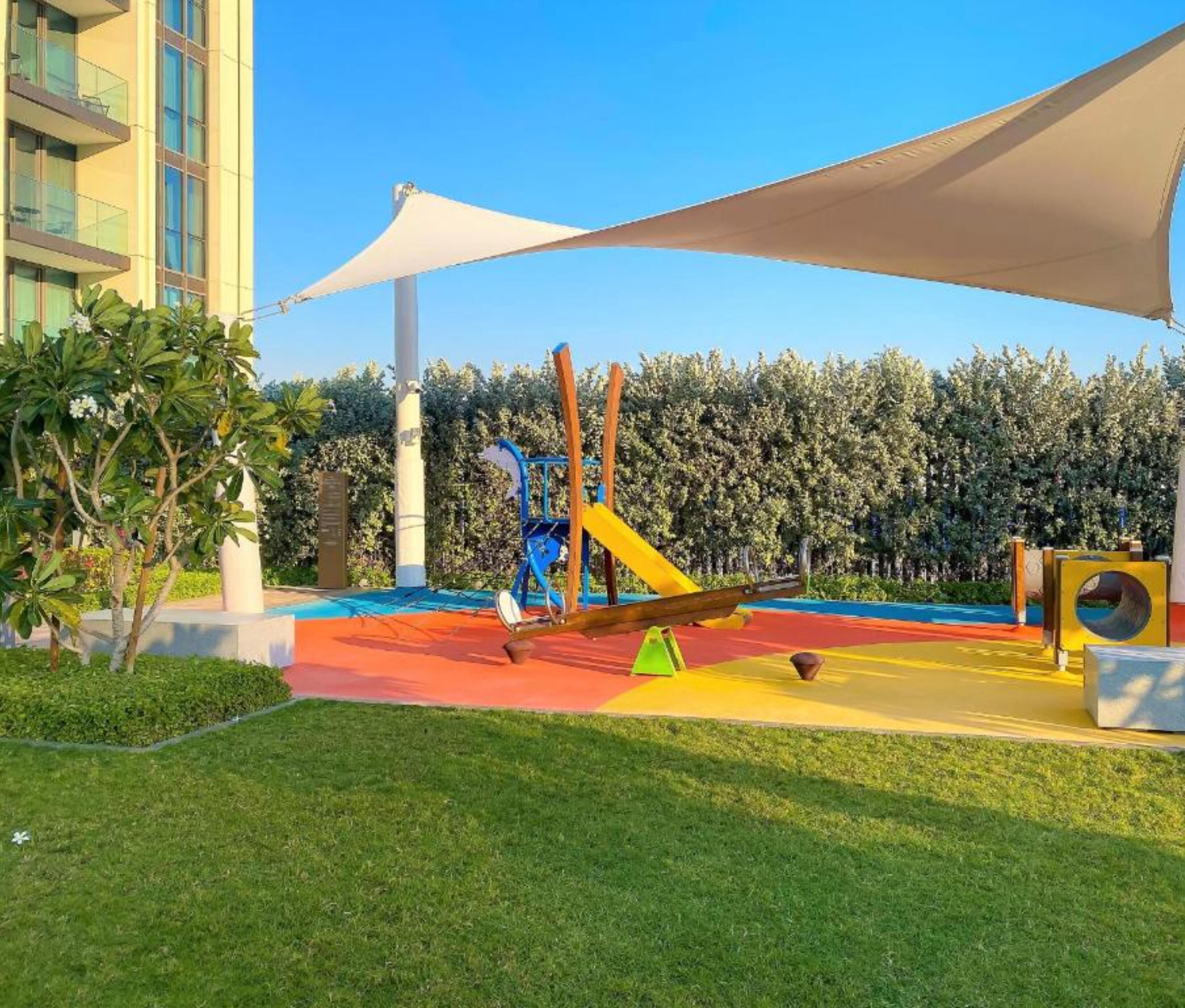 A playground with slides and swings, surrounded by a hedge and a building.