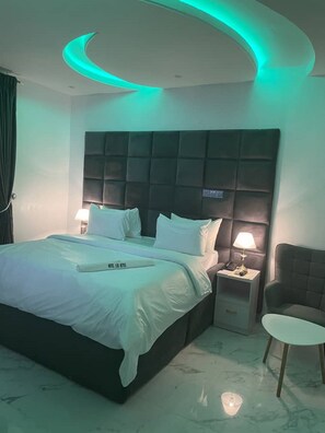 A modern bedroom with a large bed, two bedside tables with lamps, a chair, and a curved ceiling light. - HOTEL LOS REYES (Benin City)