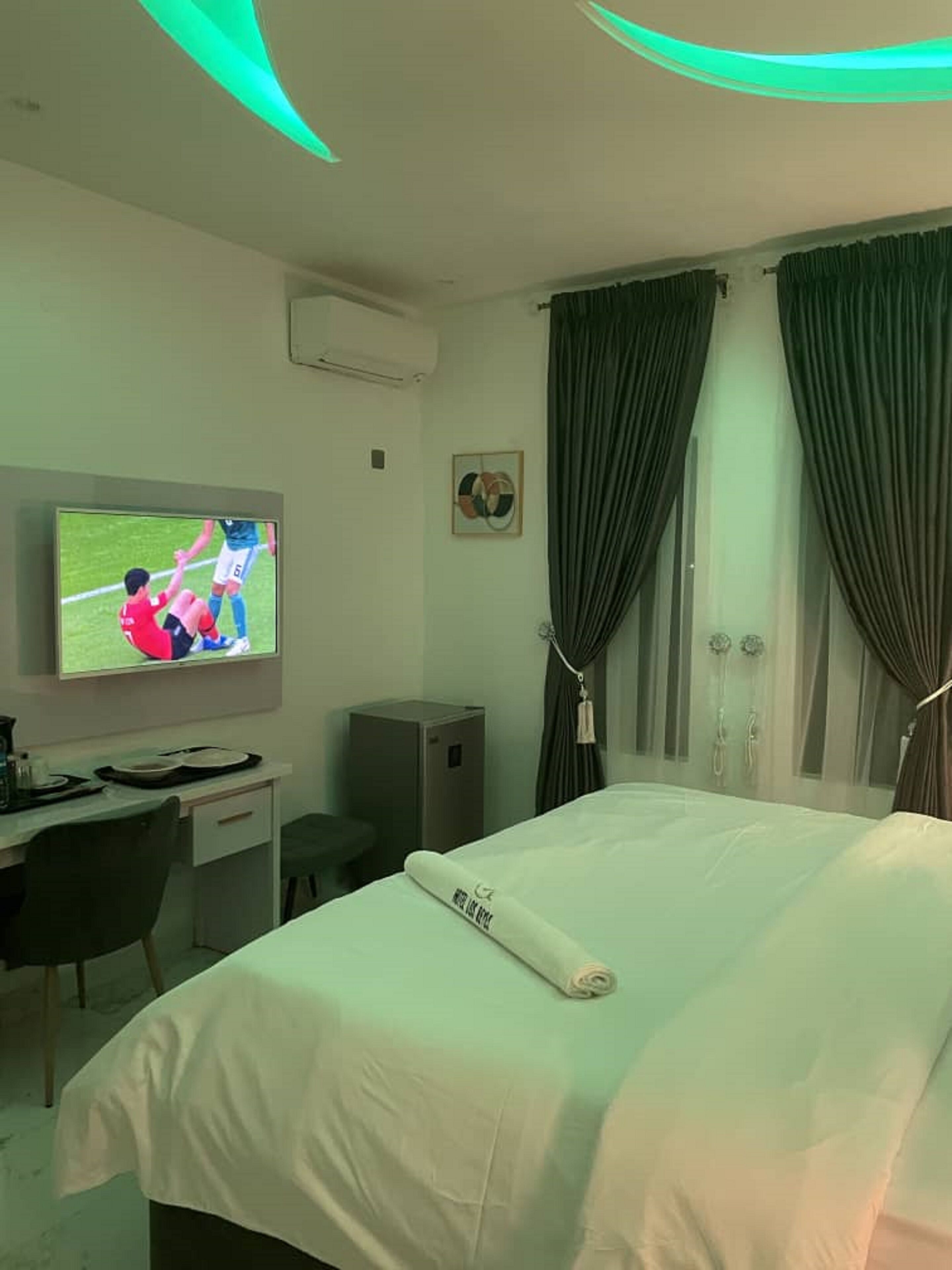 Une chambre d’hôtel avec un lit, un bureau, un téléviseur diffusant un match de football et des rideaux.