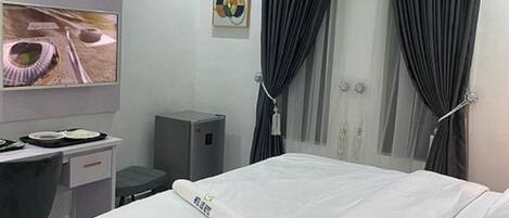 Een moderne hotelkamer met een opgemaakt bed, een bureau met stoel, een flatscreen televisie aan de muur en een raam met donkere gordijnen.