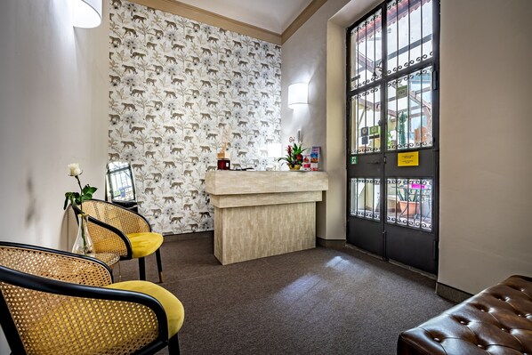 A reception area with a patterned wallpaper, a reception desk, two wicker chairs with yellow cushions, and a large window with metal bars. - Maison Pitti Florence (Florence)