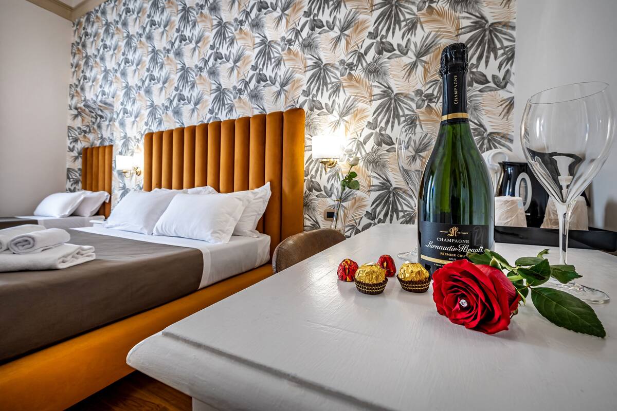 A hotel room with two beds, a table with a bottle of champagne, a glass, and a red rose.