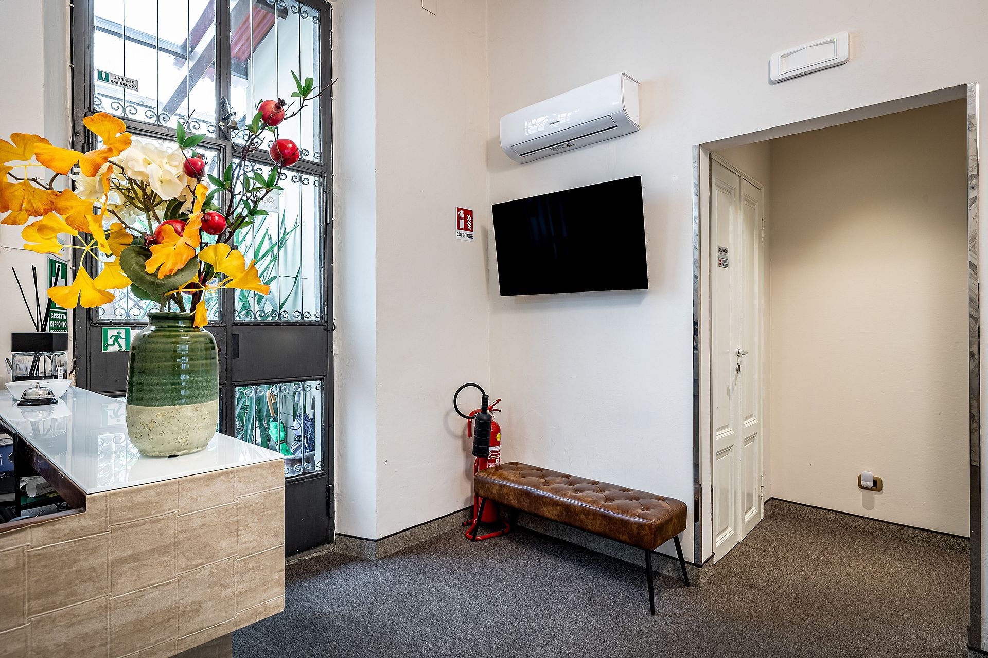 A hotel room with a large window, a flat-screen TV, a bench, and a fire extinguisher.