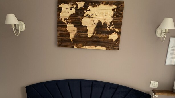 A neatly made bed with white and blue pillows, a world map artwork on the wall, and a dark blue headboard. - LE DOMAINE DU GRAVIER (Gramat)