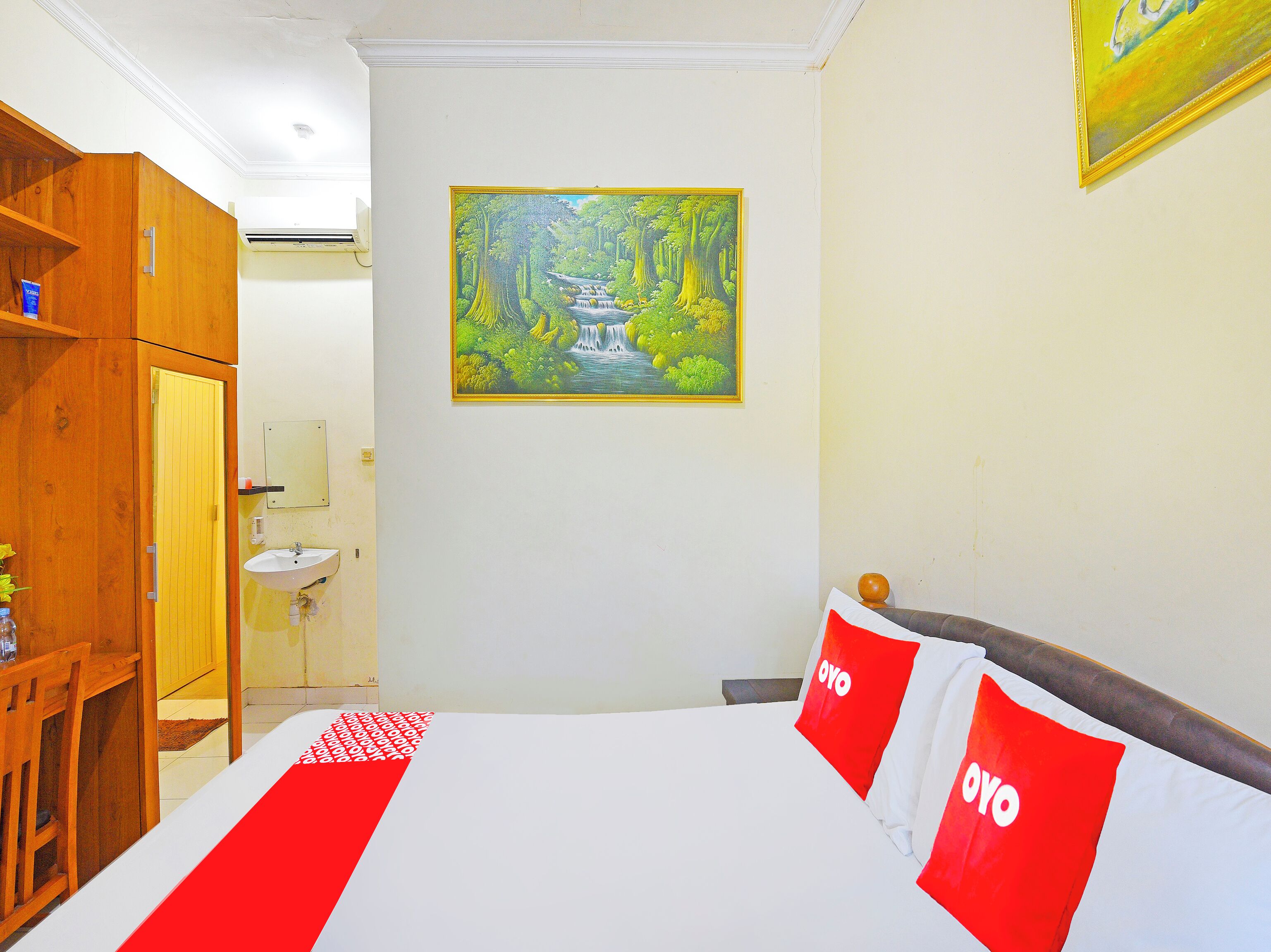Ein Hotelzimmer mit Bett, einem Holzschrank, einem Waldgemälde und einem im Hintergrund sichtbaren Badezimmer.