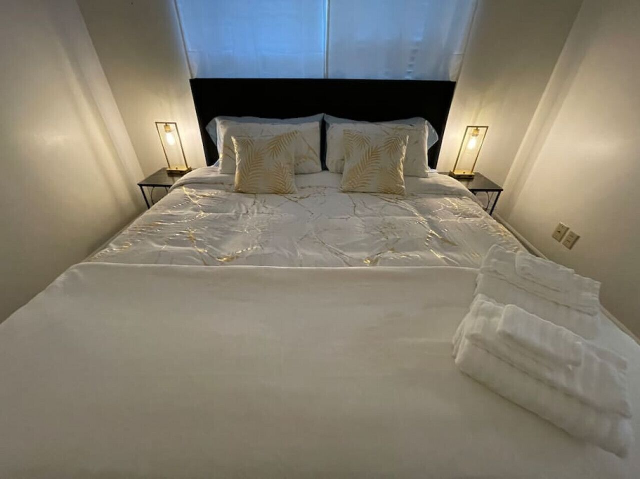 Una cama bien hecha con sábanas y almohadas blancas, dos lámparas de noche y una toalla doblada a un lado.