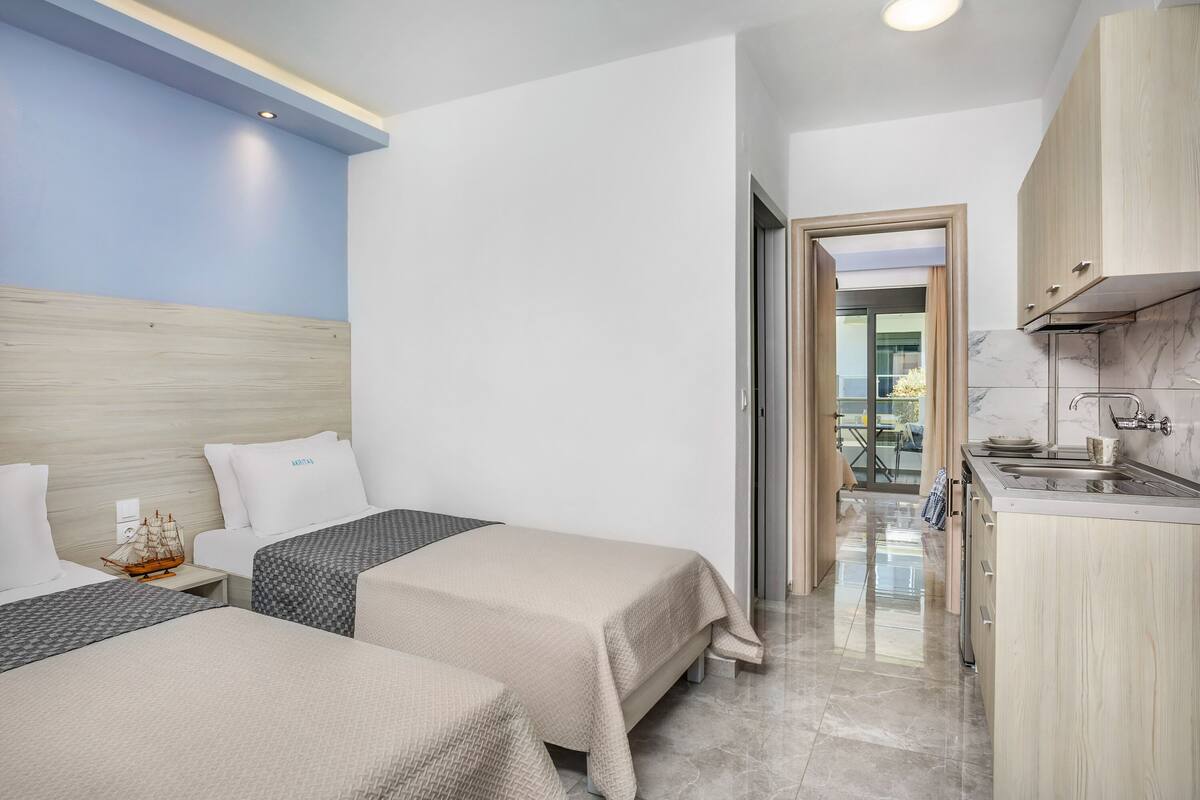 A hotel room with two beds, a small nightstand with a ship model, a kitchen area with a sink and cabinets, and a view of the outside balcony.
