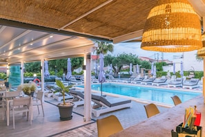 An outdoor pool area with lounge chairs and umbrellas, a covered seating area with tables, and a large hanging light fixture.