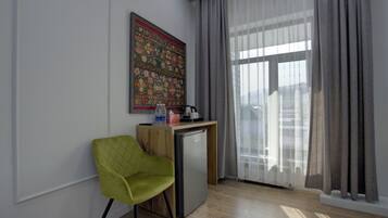 A small, modern room with a green chair, a wooden desk, a refrigerator, and a large window with sheer curtains.