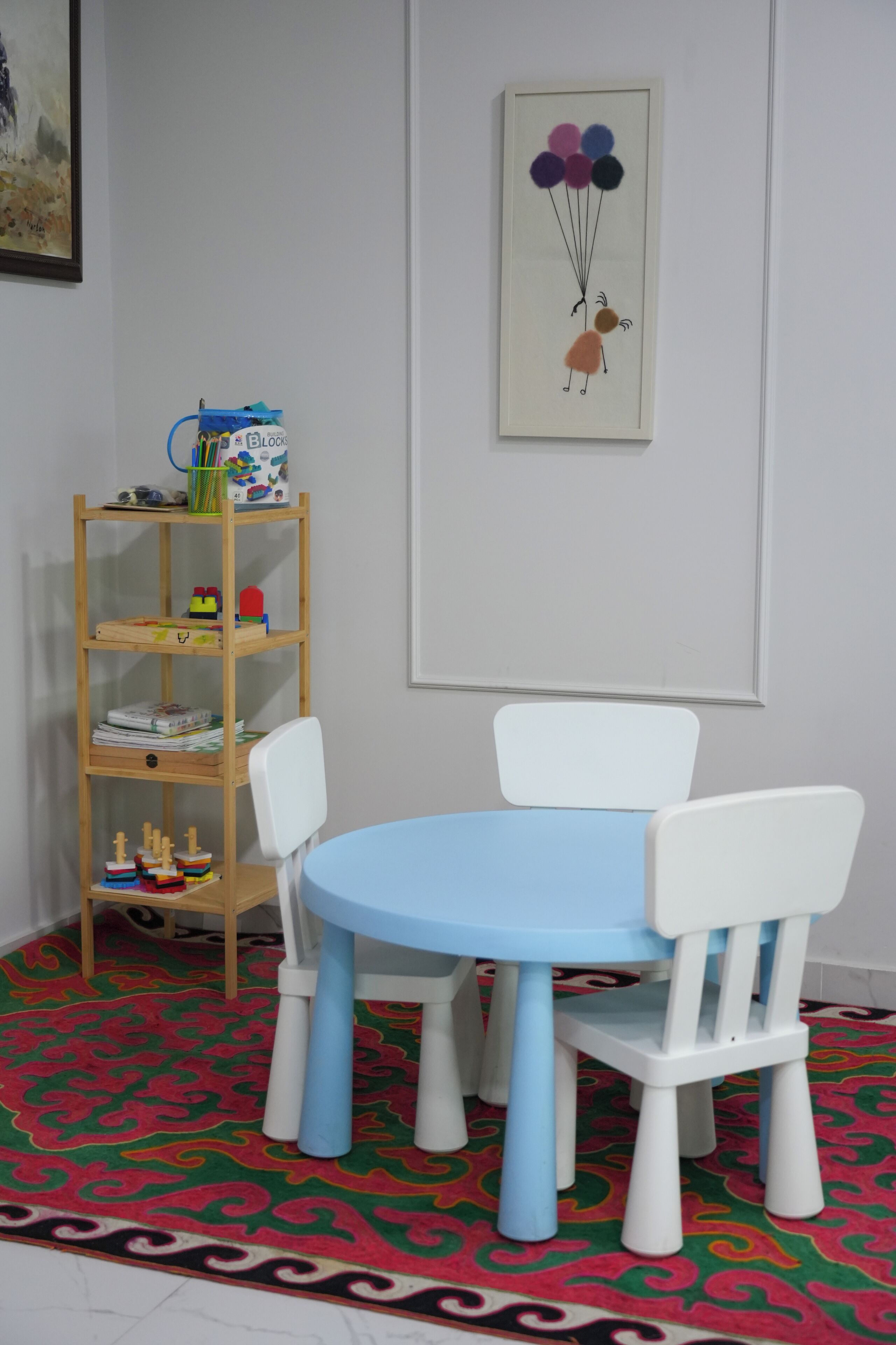 Una habitación con una mesa azul y sillas blancas, una estantería de madera con juguetes, y un cuadro con globos en la pared.