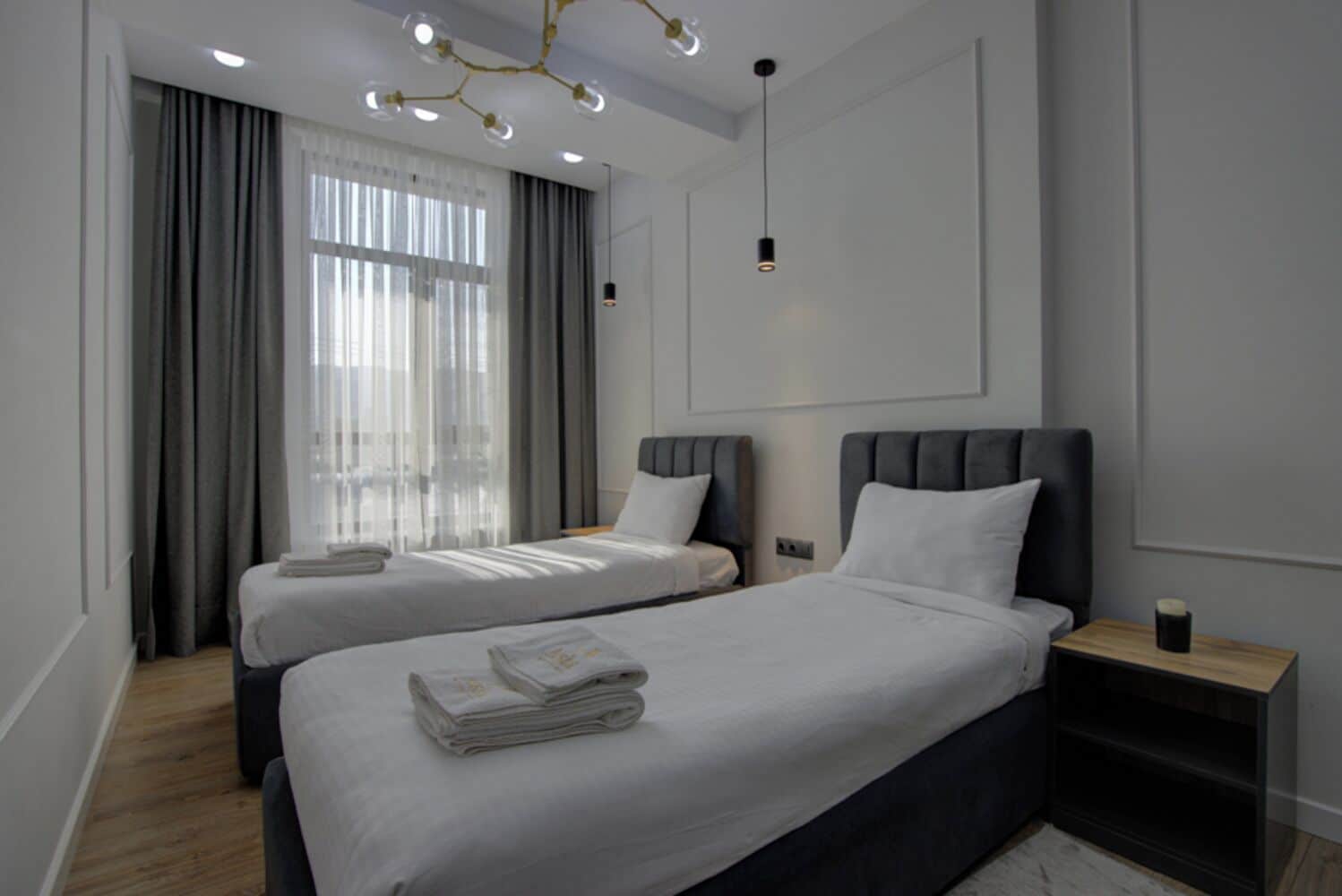 Ein modernes Hotelzimmer mit zwei Einzelbetten, einem großen Fenster mit Vorhängen, einer Kristallleuchtre und einem kleinen Nachttisch mit einer Tasse.